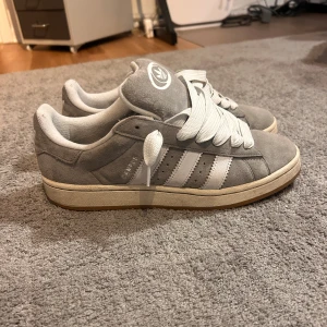 Adidas Campus grå sneakers mocka - Säljer ett par Adidas Campus sneakers i grå mocka med vita detaljer och klassiska tre ränder på sidan. Skorna har breda vita snören, rund tå och platt sula. Adidas logga på plösen och insidan. Perfekta för dig som gillar retrostil och streetwear. Äkta och typ aldrig använda.