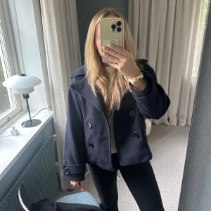 Mörkblå kappa - Säljer en mörkblå skepparkavaj med dubbelknäppning och stora svarta knappar. Jackan har bred krage och är lite kortare i modellen, vilket ger en snygg och trendig look. Perfekt för dig som gillar klassisk stil med en modern twist.