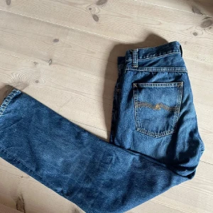 Blå jeans från Nudie Jeans W30 L30 - Snygga blå jeans från Nudie Jeans med klassisk femficksdesign och gul kontrastsöm på bakfickan. Jeansen har rak passform och är tillverkade i slitstarkt denim. Perfekta för dig som gillar en tidlös och stilren look.