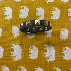 Svart armband med guldnitar - Coolt svart armband i skinnimitation med stora fyrkantiga guldnitar runt om. Justerbar knäppning med tryckknappar gör att det passar de flesta handleder. Perfekt accessoar för att ge din outfit en edgy vibe.