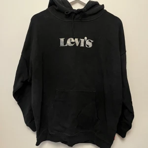 Svart hoodie från Levi's med logga - Svart hoodie från Levi's med stor vit logga på bröstet. Tröjan har huva med snörning och en stor magficka framtill. Perfekt för en avslappnad och cool stil.