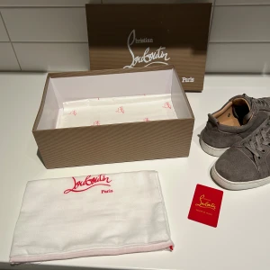 Grå mocka sneakers från Louboutin - Snygga grå sneakers från Christian Louboutin i mjuk mocka med klassisk röd sula och vita kanter. Skorna har rund tå, snörning och diskreta sömdetaljer. Insidan är fodrad i beige läder för extra komfort. Perfekta för dig som vill ha en lyxig och stilren look.