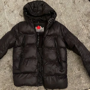 Svart pufferjacka från Zavetti Canada - Säljer en svart pufferjacka från Zavetti Canada med huva och feta loggor på både bröstet och ärmen. Jackan har dragkedjor framtill och på fickorna, samt broderad text längs insidan av huvan. Perfekt för kalla dagar när du vill hålla stilen. Storlek S.