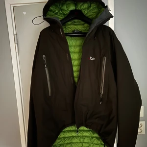 Rab dunjacka med huva, grön insida - Rab dunjacka med olivgrönt yttertyg och limegrön quiltad insida. Jackan har huva, två stora bröstfickor med dragkedja och broderad logga på bröstet. Yttermaterialet är polyamid och PTFE, fodret är dun och fjäder. Perfekt för kalla dagar och outdoor.