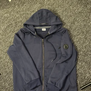Marinblå hoodie från C.P. Company - Säljer en marinblå hoodie från C.P. Company med dragkedja framtill och klassisk huva med snörning. Ärmen har den ikoniska linsen på fickan. Perfekt för dig som gillar streetwear och vill ha något stilrent men ändå unikt.