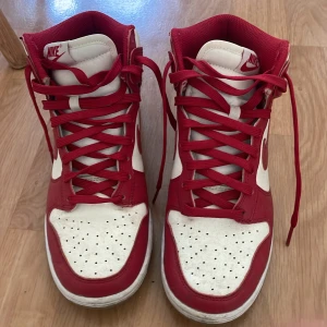 Nike Dunk High röd/vit sneakers - Nike Dunk High sneakers i rött och vitt med klassisk design. Skorna har högt skaft, röd swoosh, röda snören och perforerad tå. Ovandel i skinn och textil, yttersula i gummi. Perfekt för dig som gillar streetwear och vill sticka ut med en ikonisk modell. 