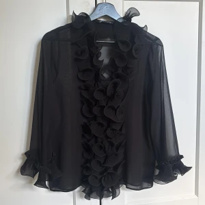 Svart volangblus från Zara XS - Säljer en svart transparent blus från Zara i storlek XS. Blusen har långa ärmar och är dekorerad med stora volanger framtill och vid ärmsluten. Tunn och luftig med en elegant och unik look, perfekt för dig som gillar statement-detaljer.