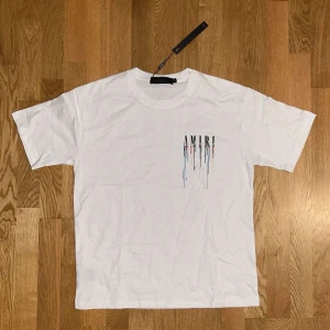 Vit AMIRI t-shirt med färgstänk - Snygg vit t-shirt från AMIRI med coolt tryck av loggan och färgstänk både fram och bak. Klassisk passform och rund halsringning. Perfekt för dig som vill sticka ut med en streetstyle-look. Tillverkad i mjuk bomull för skön känsla.