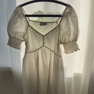 Vit kortklänning med puffärm från Vila - Supersöt vit kortklänning från Vila med puffiga korta ärmar och smockdetaljer vid ärmsluten. Klänningen har v-ringning med små knappar framtill och är gjord i ett lätt bomullstyg. Perfekt för en somrig look med romantiska vibes.