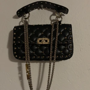 Svart väska med nitar  - Säljer en svart quiltad handväska från Val****** Gara**** med guldfärgade nitar, Väskan är väl använd med några skavanker, tappat färg på vissa ställen och tappat några nitar annars i fullt fungerande skick, priset är diskuterbart men väl sänkt redan pågrund av slitage 