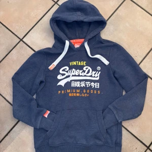 Blå Superdry hoodie med tryck - Snygg blå hoodie från Superdry med vit och gul logga samt japanska tecken på bröstet. Tröjan har huva med vita snören, känguruficka och ribbade muddar. Perfekt för dig som gillar streetwear och vill ha en bekväm och stilren look. Storlek L men sitter som en M 