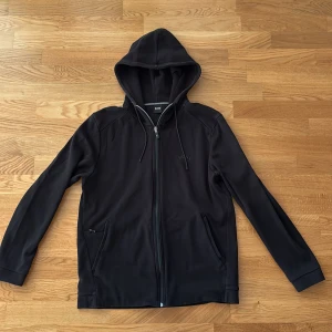 Svart hoodie från BOSS med dragkedja - Svart hoodie från BOSS med hel dragkedja framtill, justerbar huva med snören och diskret logga på bröstet. Tröjan har två fickor och är tillverkad i mjuk bomullsmix. Perfekt för en chill och stilren look.