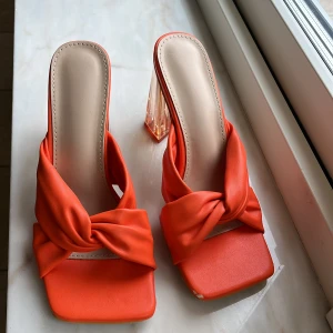 Orange sandaler med transparent klack - Snygga orange sandaler med fyrkantig tå och bred knutdetalj framtill. Skorna har en hög, transparent klack som ger en cool och modern vibe. Ovandelen är i mjukt syntetmaterial och innersulan är beige. Perfekta för dig som vill sticka ut med färg och stil.
