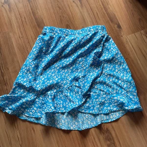 Blommig blå kjol med volang - Säljer en söt blå kjol med vitt blommönster och volangdetaljer. Kjolen har en kort längd och är perfekt för varma dagar. Materialet känns lätt och luftigt, vilket gör den skön att bära. Fräsch och trendig stil för dig som gillar färg och mönster.