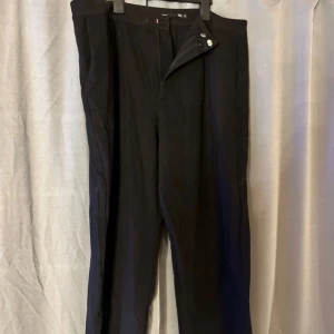 Svarta kostymbyxor från 157, XL - Svarta kostymbyxor från 157 i storlek XL. Klassisk rak passform med diskret knapp- och dragkedjestängning framtill. Snygga att styla med skjorta eller t-shirt. Materialet känns mjukt och har en lätt struktur.