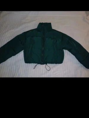 Mörkgrön croppad pufferjacka - Snygg mörkgrön croppad pufferjacka med hög krage och dragkedja framtill. Jackan har snörning i nederkanten för justerbar passform och långa ärmar. Perfekt för dig som vill ha en trendig och varm jacka till höst och vinter.