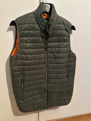 Mörkgrön quiltad dunväst - Stilren mörkgrön quiltad väst med hög krage och hel dragkedja framtill. Västen har två praktiska fickor med dragkedja och en orange insida som ger en snygg kontrast. Perfekt för lager-på-lager. Strl S.