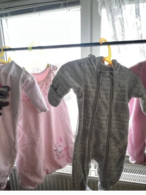 Babysoveraller och pyjamasar i pastell 68/74 - Tre mysiga babyplagg: en grå overall med huva och dragkedja i mjukt mönstrat tyg, en rosa sovpåse med broderad nalle och hjärtan samt en ljusrosa pyjamas med gulligt djurmotiv. Perfekta för sköna hemmakvällar och sovstunder. Alla i mjuka material.