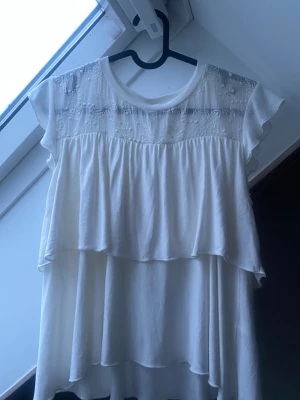 Vit blus/Biała koszula - Romantisk vit blus med korta ärmar, detaljerad spets upptill och blommigt broderi. Blusen har dubbla volanger som ger ett flowy och luftigt intryck samt rund halsringning. Perfekt för dig som gillar en feminin och söt stil.