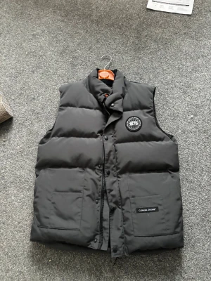 Grå dunväst från Canada Goose - Snygg grå dunväst från Canada Goose med hög krage och två stora framfickor. Västen har ett klassiskt Canada Goose-märke på bröstet och stängs med både dragkedja och knappar. Perfekt för lager-på-lager och håller dig varm under kyliga dagar.
