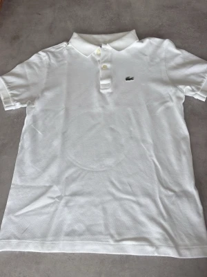 Vit pikétröja från Lacoste - Klassisk vit pikétröja från Lacoste med korta ärmar och liten broderad logga på bröstet. Tröjan har krage och knappar framtill. Perfekt till en clean och stilren look.