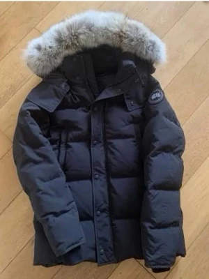 Canada Goose Wyndham Black Label - Säljer en svart dunjacka från Canada Goose med pälskantad huva och klassisk patch på ärmen. Jackan är quiltad, har lång ärm och dragkedja framtill. Perfekt för kalla vinterdagar och riktigt snygg streetstil.