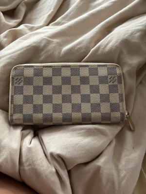 Beige-rutig Louis Vuitton plånbok - Snygg beige och grå-rutig plånbok från Louis Vuitton med dragkedja runtom. Plånboken har flera fack för kort och sedlar samt ett myntfack med dragkedja. Klassisk Damier-mönster och detaljer i ljust skinn. Perfekt för dig som gillar lyxiga accessoarer. Orginalpris - 9000-10,000