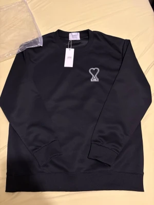 Svart sweatshirt från Ami Paris - Säljer en svart sweatshirt från Ami Paris med det ikoniska hjärta-A broderiet på bröstet. Tröjan har rund hals, ribbade muddar och är tillverkad i mjukt bomullsmaterial. Perfekt för en clean och stilren look.