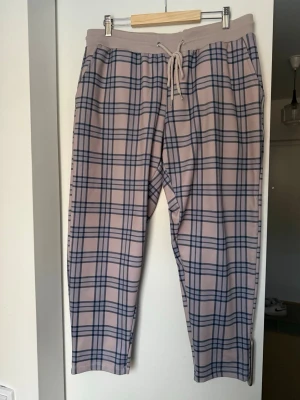 Beige rutiga mjukisbyxor med snörning - Säljer ett par beige mjukisbyxor med mörkblått rutmönster och snörning i midjan. Byxorna har avslappnad passform och resår i midjan för extra komfort. Perfekta för chill dagar eller när du vill vara extra bekväm.