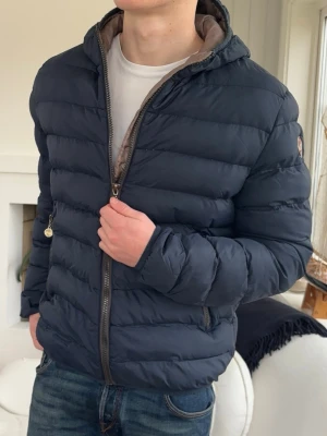 Mörkblå dunjacka med huva - Snygg mörkblå dunjacka med huva och dragkedja framtill. Jackan har quiltad design och två sidofickor med dragkedja. Perfekt för kyliga dagar och enkel att matcha till olika outfits.