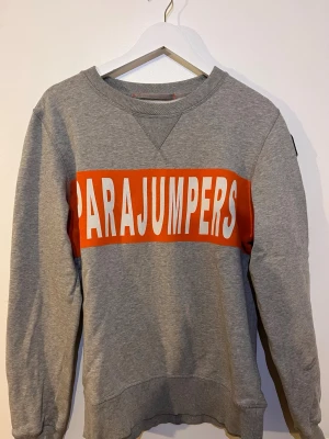 Grå sweatshirt från Parajumpers - Snygg grå sweatshirt från Parajumpers med stor logga i vitt på en orange panel över bröstet. Tröjan har rund halsringning, ribbade muddar vid ärmslut och nederkant samt en avslappnad passform. Perfekt för en casual och sportig stil.