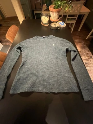 Blå stickad tröja från Polo Ralph Lauren - Snygg blå stickad tröja från Polo Ralph Lauren med klassisk rund halsringning och diskret broderad logga på bröstet. Tröjan har lång ärm och är gjord i ett mjukt, strukturerat material som känns skönt mot huden. Perfekt för dig som gillar stilrena plagg.