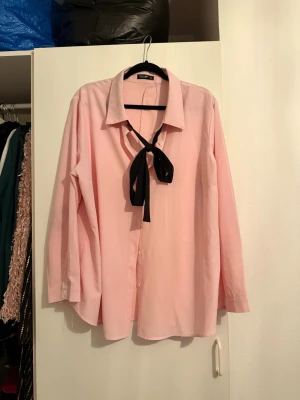 Ljusrosa skjorta med svart knytband - Säljer en ljusrosa skjorta från Zara med klassisk krage och svart knytband vid halsen. Skjortan har lång ärm och knäppning framtill. Perfekt för dig som vill ha en stilren men ändå unik look.