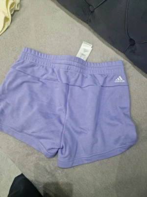 Ljuslila Adidas shorts med snörning - Säljer ett par ljuslila shorts från Adidas med vit logga på ena benet och snörning i midjan. De har en sportig look och är perfekta för träning eller chill. Tillverkade i mjukt material som känns skönt mot huden.