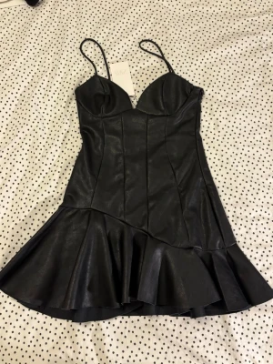 Svart klänning i skinnimitation Zara - Supersnygg svart kort klänning från Zara i skinnimitation med tunna axelband och volangkant nedtill. Helt ny med etikett kvar!