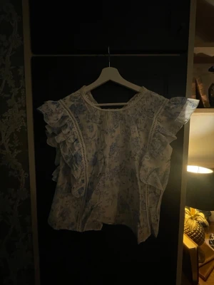 Blommig volangblus i blått och vitt - Supersöt blus med blått blommönster på vit botten. Blusen har volangdetaljer längs axlarna, spets vid halsringningen och en liten knapp i nacken. Knäppning i ryggen med en knapp. Är kort för mig som är 179 cm.