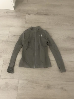 Grå träningsjacka från Xshape - Slimmad grå träningsjacka från Xshape med hel dragkedja framtill och tumhål i ärmsluten. Jackan har hög krage och diskret logga på bröstet. Tillverkad i ett stretchigt funktionsmaterial som andas, perfekt för träning och aktiva dagar.Dock ett litet håll i vänster under arm
