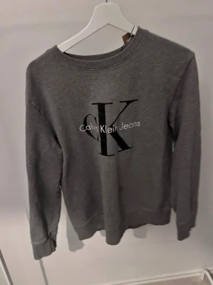 Grå sweatshirt från Calvin Klein - Snygg grå sweatshirt från Calvin Klein Jeans med stort svart CK-tryck och vit text på bröstet. Tröjan har rund halsringning, ribbade muddar vid ärmslut och nederkant. Perfekt för en avslappnad och trendig look.