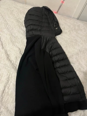 Svart dunjacka med huva från Moncler - Snygg svart dunjacka från Moncler med quiltade partier och stickade ärmar. Jackan har en stor huva och dragkedja framtill. Klassisk Moncler-logga på ärmen och en modern, sportig look som passar perfekt till vardags.