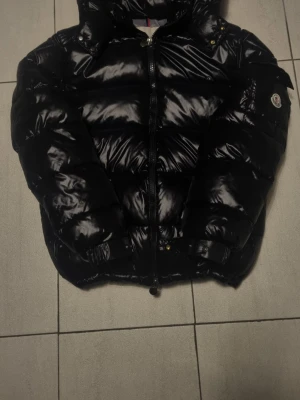 Monster maya - Säljer en svart glansig dunjacka från Moncler med klassisk pufferdesign. Jackan har huva, dragkedja framtill och Moncler-logga på ärmen. Perfekt för kalla dagar och riktigt snygg till streetwear-stilen.