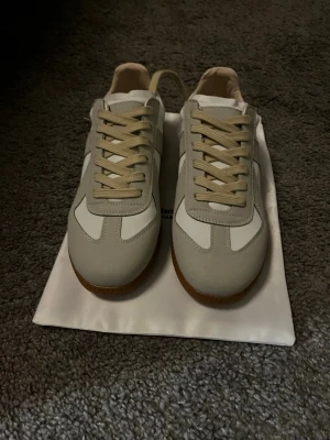 Vita och beige sneakers med mockadetaljer - Snygga sneakers från Maison Margiela i vitt och beige med klassiska paneler i mocka och skinn. Skorna har beige snörning och en brun gummisula som ger en retrovibe. Perfekta för dig som gillar stilrena och trendiga sneakers. Kom privat för mer info!!!