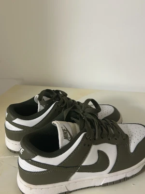 Nike Dunk Low i vitt och olivgrönt - Nike Dunk Low sneakers i vitt och olivgrönt läder med klassisk swoosh på sidorna. Skorna har snörning, perforerad tå och låg profil. Perfekta för dig som gillar streetstyle och vill ha en clean look. Knappt använda ser ut som nya, priset kan diskuteras vid snabb affär pågrund av flytt 