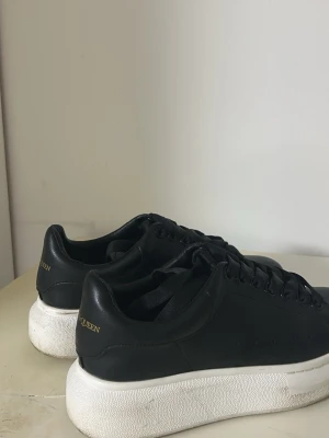 Svarta Alexander McQueen sneakers - Stilrena svarta sneakers från Alexander McQueen med vit, chunky platt sula och guldfärgad logga på hälen. Skorna har snörning och är tillverkade i skinn för en lyxig känsla. Perfekta för dig som gillar minimalistisk och modern design. Priset kan diskuteras vid snabb affär pågrund av flytt. 