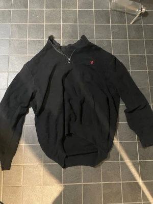 Svart halvzip från Ralph Lauren XL - Svart långärmad tröja från Ralph Lauren med halv dragkedja och röd broderad logga på bröstet. Tröjan har en klassisk passform och ribbade muddar vid ärmslut och midja. Perfekt för en avslappnad och stilren look.