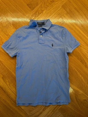 Blå pikétröja från Polo Ralph Lauren - Klassisk ljusblå pikétröja från Polo Ralph Lauren med krage, knappar och broderad logga på bröstet. Perfekt för en avslappnad men stilren look.