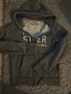 Grå zip-hoodie från Hollister - Mysig grå hoodie från Hollister med dragkedja och vit text på bröstet. Modellen har huva med dragsko, två fickor fram och ribbade muddar vid ärmslut och nederkant. Perfekt för en avslappnad och stilren look. 😁 Hör av vid intresse
