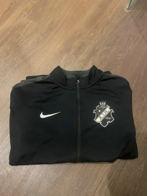 Svart Nike AIK träningströja - Svart träningströja från Nike med AIK:s klubbmärke på bröstet och vit Nike-logga. Tröjan har hel dragkedja, ståkrage och två sidofickor. Tillverkad i ett lätt och funktionellt material som passar perfekt till träning. Mer frågor kom privat😃