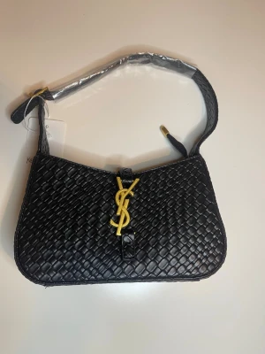 Svart axelväska från Yves Saint Laurent - Snygg svart axelväska från Yves Saint Laurent i flätat skinn med guldigt YSL-emblem framtill. Väskan har en kort axelrem och elegant formad siluett. Perfekt för dig som vill ha en stilren och lyxig accessoar med exklusiv känsla.