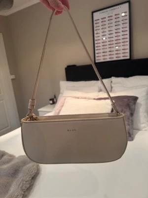 Beige handväska från NA-KD - Stilren beige handväska från NA-KD med minimalistisk design och guldfärgade detaljer. Väskan har en avlång, rundad form och justerbar axelrem. Perfekt för att lyfta din outfit med en clean och modern touch.