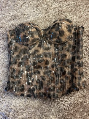 Leopardmönstrad paljettkorsett - Snygg korsett med leopardmönster och glittriga paljetter över hela plagget. Kupor med bygel för extra stöd och figurnära passform. Insida i beige tyg. Perfekt för dig som vill sticka ut och glänsa.